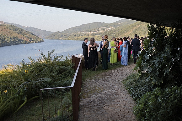Wedding cocktail hour with guests mingling in formalwear, holding cocktails and wearing sunglasses on a lakeside terrace with mountain views