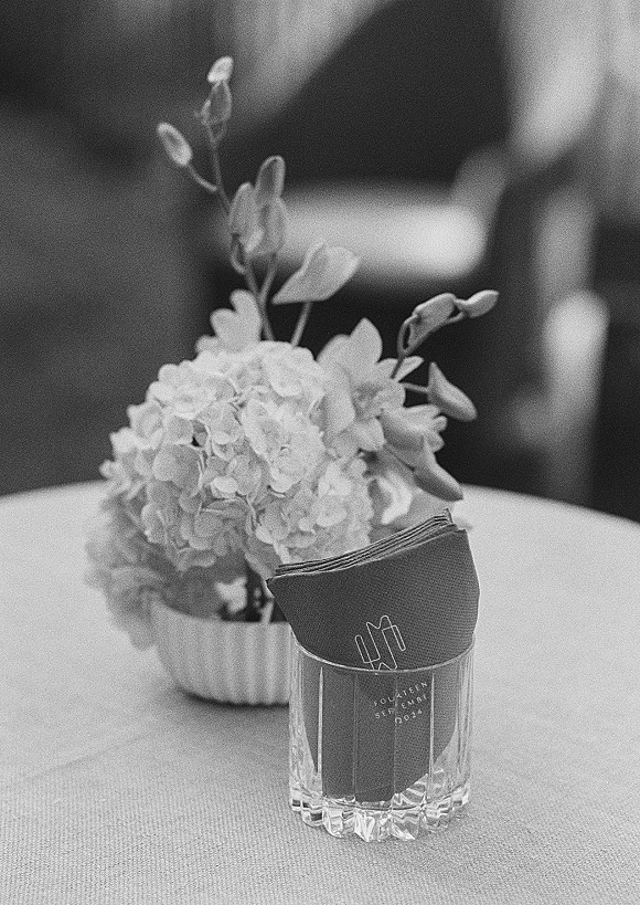 Reception table decor with wedding cocktail napkins in a crystal tumbler beside hydrangea bud vase and folded napkins in an indoor reception space