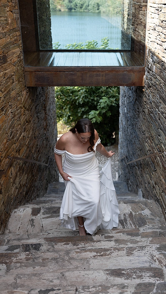 Bridal portrait of a bride walking up stairs in an off the shoulder wedding dress, lifting her satin train on a stone staircase by a lake