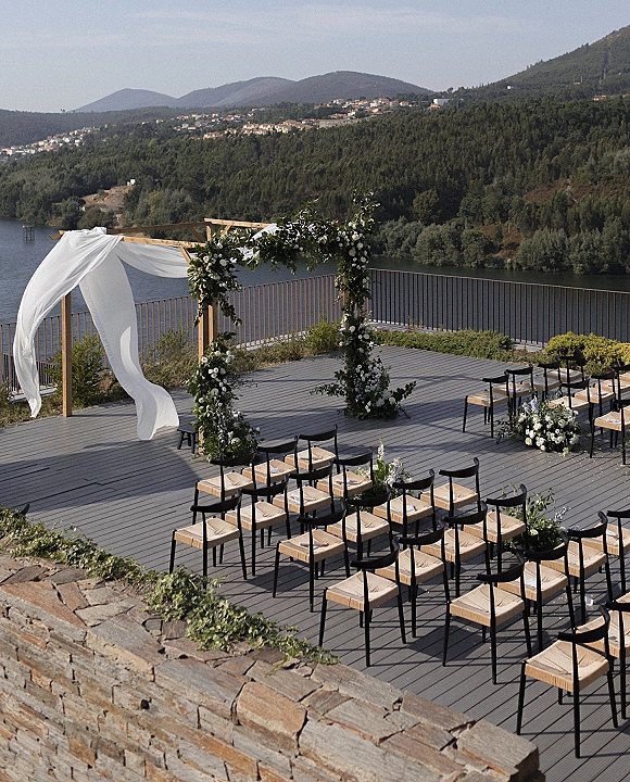 Ceremony setup with outdoor ceremony seating facing a floral arch draped in white fabric on a lakeside terrace with mountain hills