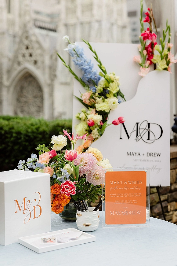 Wedding welcome sign on an acrylic wedding sign beside a blue delphinium arrangement at an outdoor courtyard guestbook table by stone walls