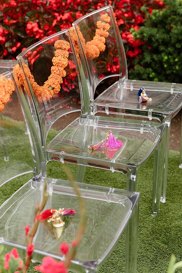 Wedding ceremony chairs in clear acrylic with orange marigold garlands, tassel favors and mini figurines set on a green garden lawn