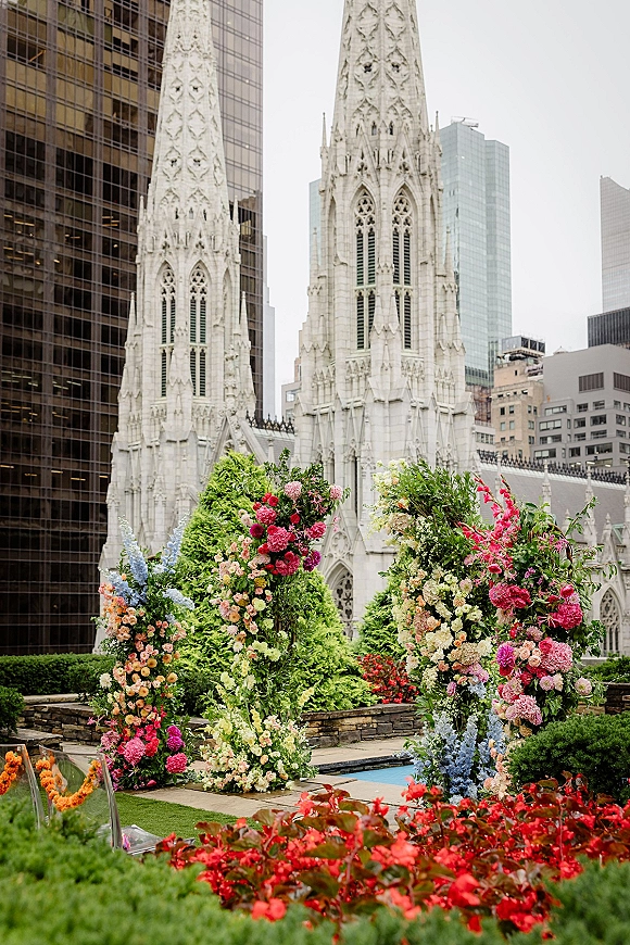 Ceremony floral arch in an asymmetrical floral arch design with mixed blooms and greenery by a pool, cathedral spires beyond