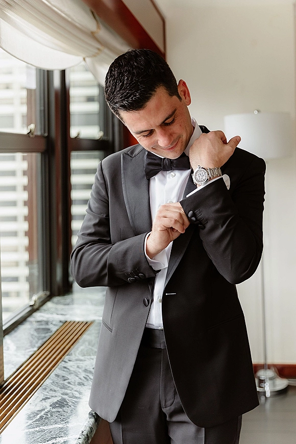 Groom portrait in a black tuxedo adjusting cufflinks by a window, bow tie and wristwatch visible with city buildings beyond curtains