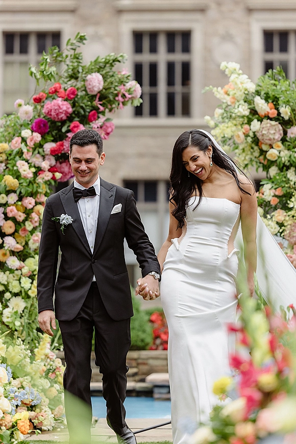 Couple portrait of bride and groom walking hand in hand under a colorful floral arch on a garden walkway by a stone building