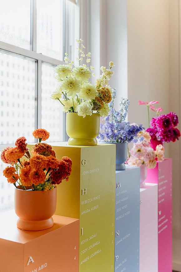 Wedding seating chart on colorful pedestals with pastel floral arrangements in ceramic vases, set by large windows with city views