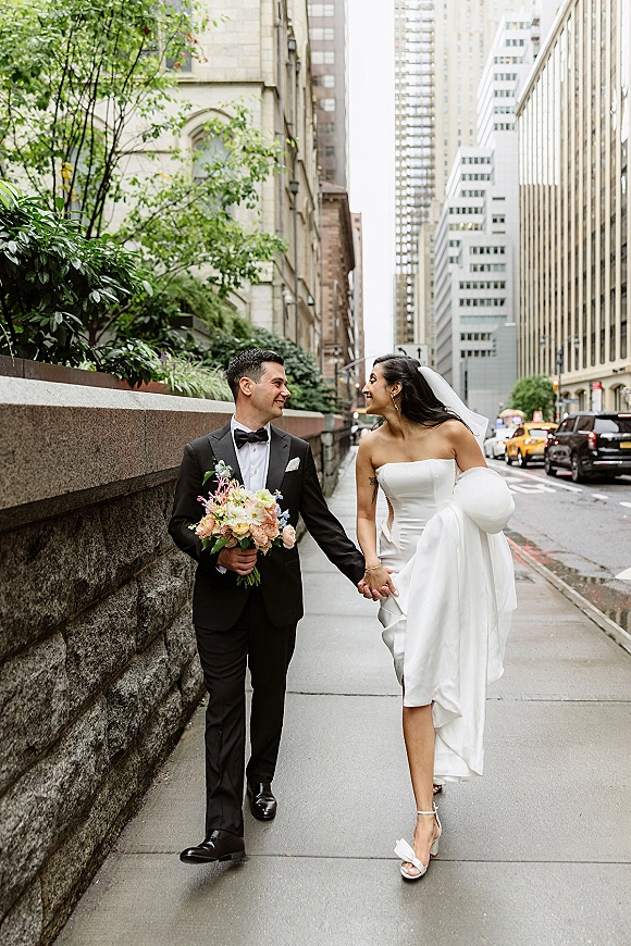 Couple portrait of bride and groom walking hand in hand on a city sidewalk, bride holding bouquet and dress with veil trailing