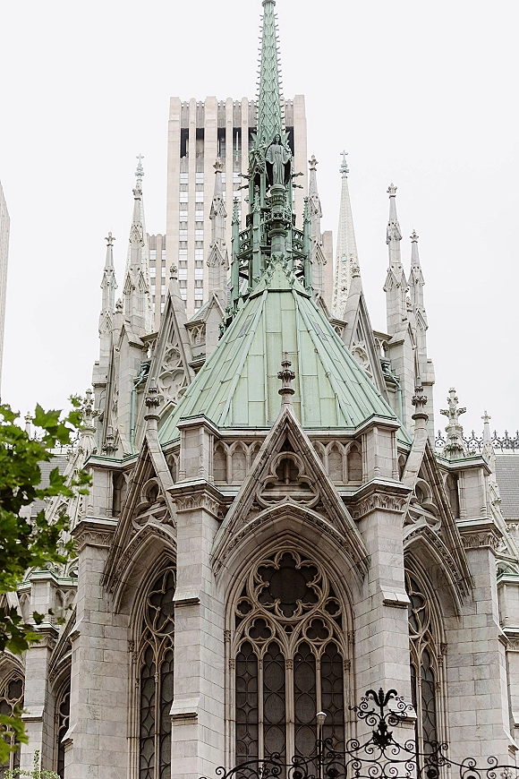 Church exterior with gothic church architecture, stone spires and a copper dome behind a wrought iron fence against city buildings and trees