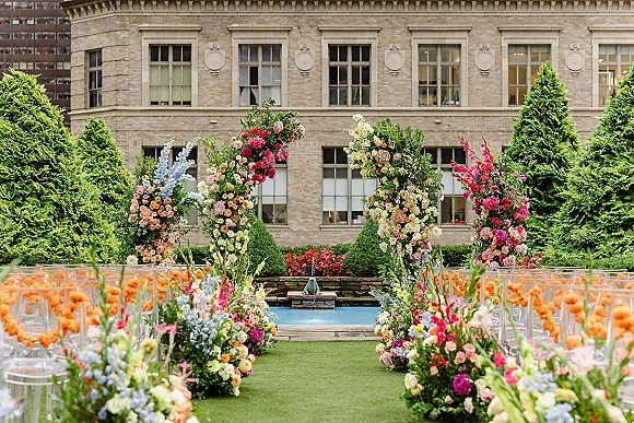 Ceremony aisle decor with floral wedding arch, orange garlands and greenery lining a grass aisle with clear chairs near a fountain