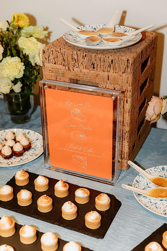 Wedding dessert display with mini tarts and custard spoons on patterned plates, slate tray, acrylic sign, and floral accents on linen tablecloth