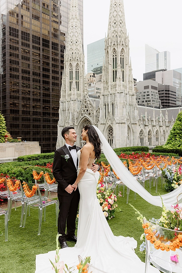 Couple portrait of bride and groom embracing in aisle, veil train flowing, by clear acrylic chairs and bright florals on terrace lawn