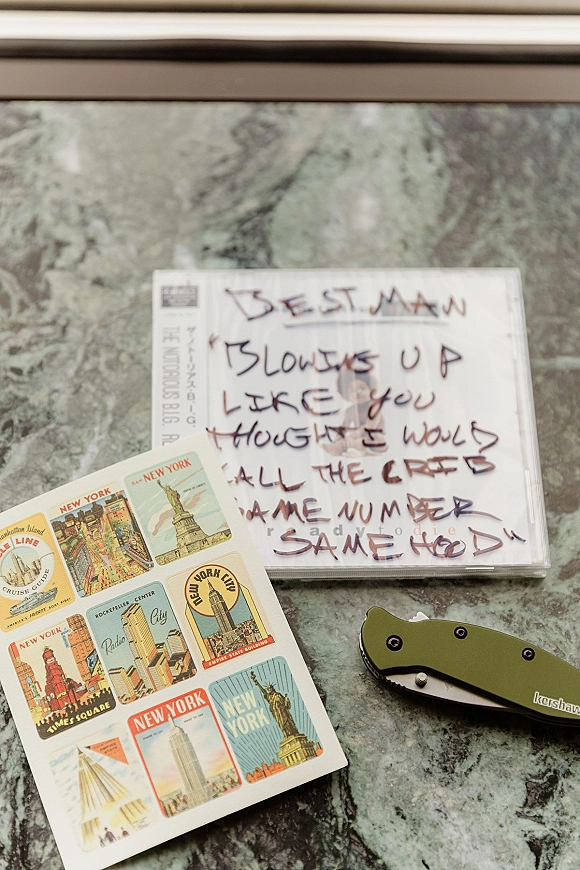 Wedding details flat lay with getting ready details, including a CD case, handwritten note, postcard stickers and pocket knife on marble surface