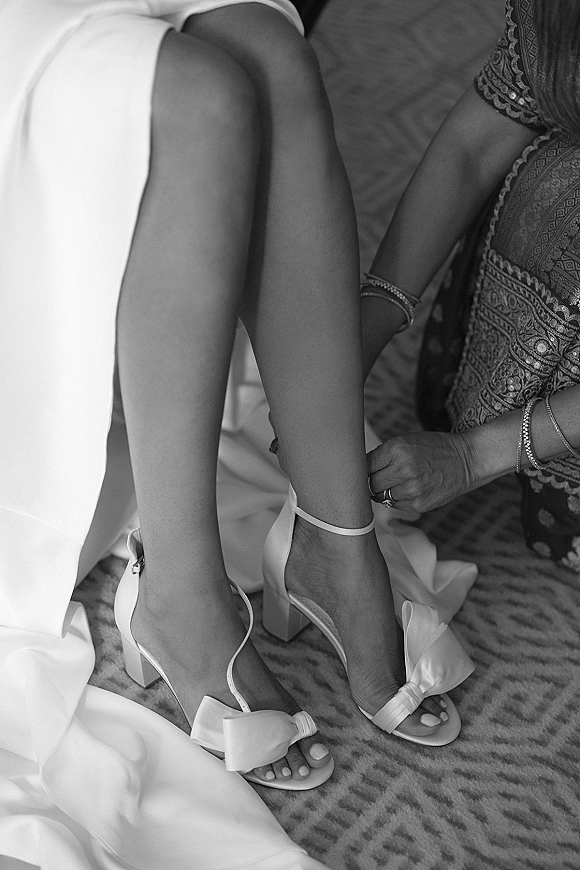 Wedding shoes and bridal heels with ankle strap and bow as a bride fastens them, jewelry and gown hem visible on a carpeted floor