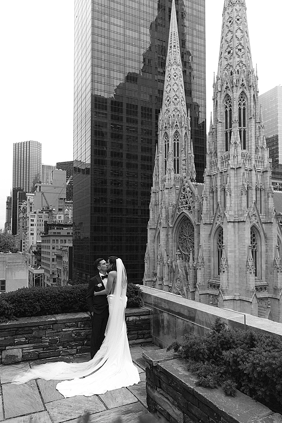 Wedding kiss portrait in black and white, bride with long veil and train kissing groom in tuxedo on a rooftop terrace by cathedral spires