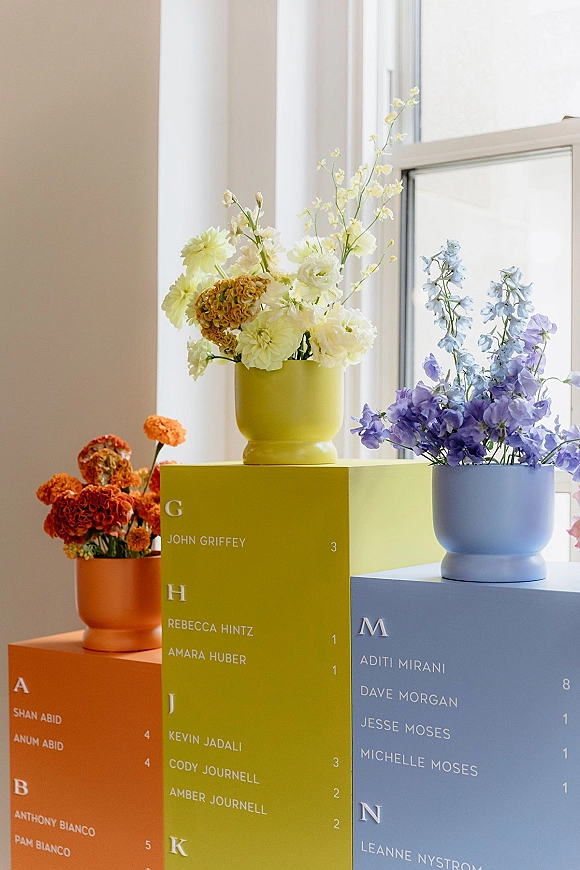 Wedding escort cards in a color block alphabetical escort card display on pedestal boxes, with flower vases by a window in natural light