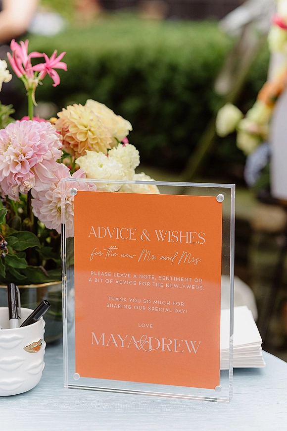 Wedding advice sign in a clear acrylic frame with coral lettering, pens and advice cards beside dahlias and greenery on linen table outdoors