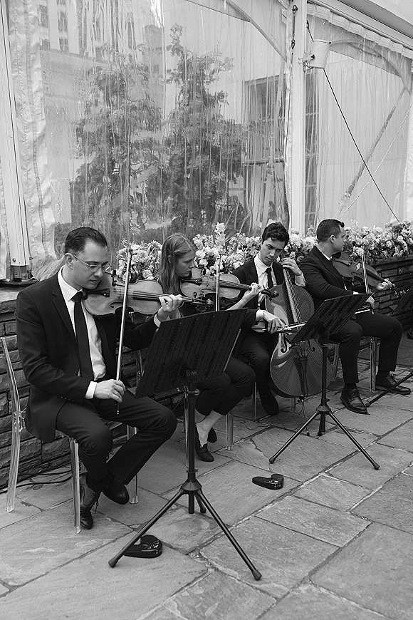 Wedding string quartet playing live wedding music under a clear-walled tent, with violins and cello on a stone patio by trees