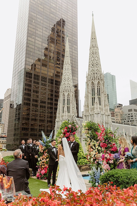 Wedding ceremony on a rooftop terrace with bride and groom at the floral arch altar, city skyline and church spires behind guests