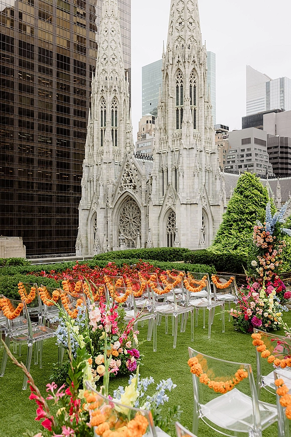 Ceremony setup for a rooftop wedding ceremony with clear acrylic chairs, marigold garlands, and a floral arch before cathedral and skyline