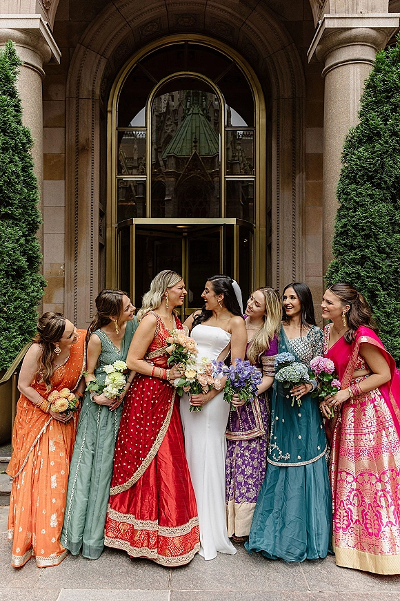 Bridesmaids group photo with bride with bridesmaids, laughing in colorful lehengas holding bouquets outside a stone arched entrance with veil