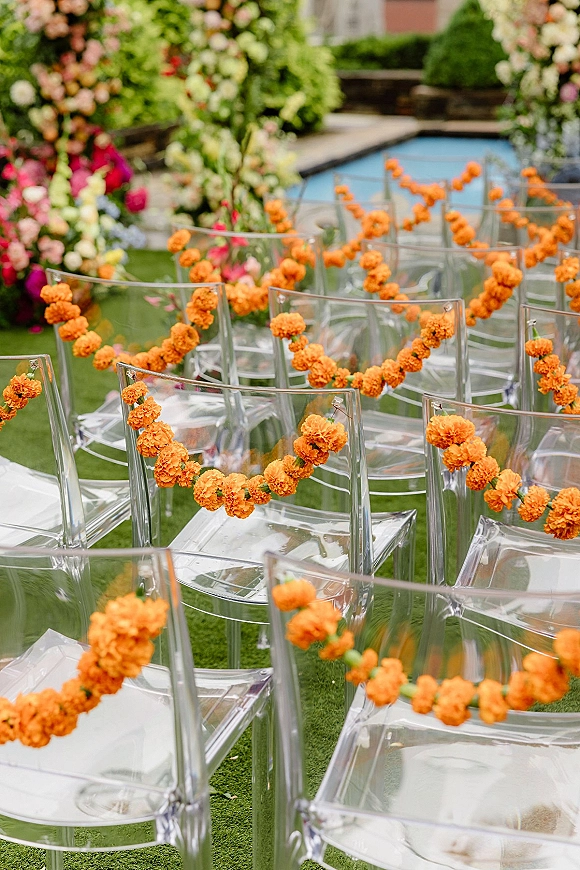 Ceremony seating decor with clear acrylic wedding chairs lined along a garden aisle, draped in orange marigold garlands and florals on grass lawn