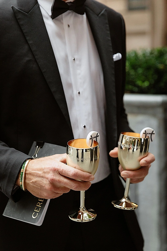 Wedding cocktail hour drinks served in silver goblet cups by a tuxedoed groom with bow tie, outdoors near greenery and stone planter