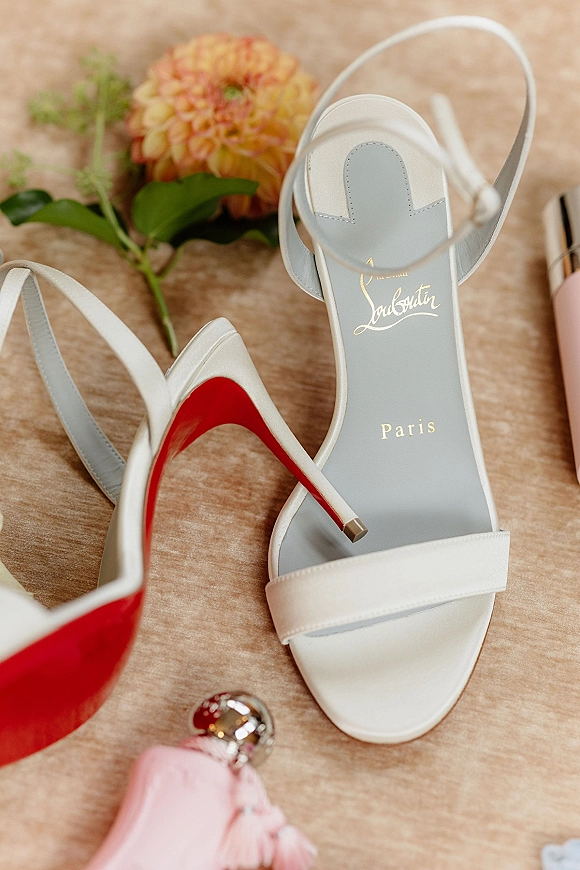 Bridal shoes flatlay of white strappy wedding heels with red soles, orange flower, greenery, perfume and lipstick on a wood tabletop