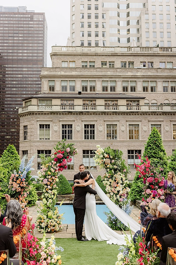 Ceremony kiss as the bride and groom embrace under a colorful floral arch, her veil flowing on a rooftop terrace with city skyline behind