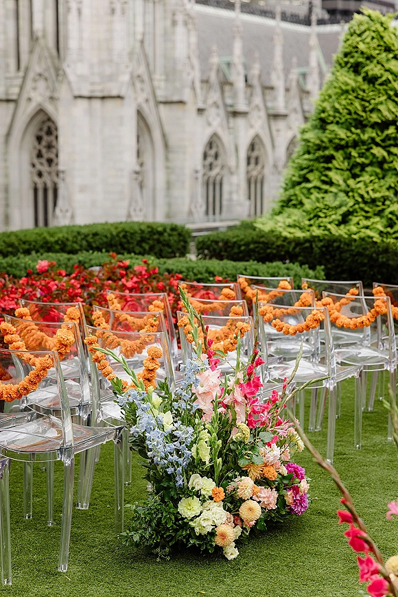 Ceremony seating with clear acrylic wedding chairs lined in rows, marigold garlands draped along the aisle before a stone cathedral facade