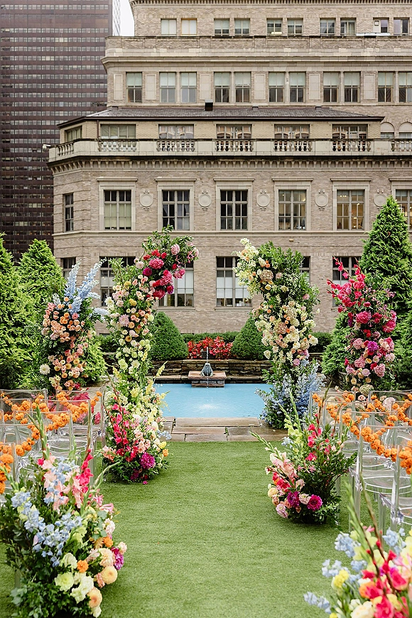 Ceremony aisle decor with floral ceremony arch of colorful wedding flowers, clear acrylic chairs, and grass runner in courtyard by fountain