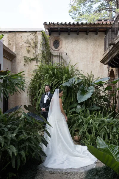 Couple portrait of bride with cathedral veil and long dress train beside groom in tuxedo, framed by lush courtyard greenery and brick archway