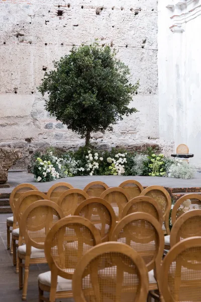 Ceremony setup with rattan wedding chairs facing a low white-and-green floral ground arrangement on a platform in a stone-walled courtyard