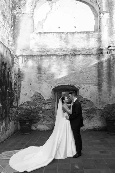 Wedding couple portrait in black and white, bride in strapless gown and long veil touching groom’s face by an arched stone doorway courtyard