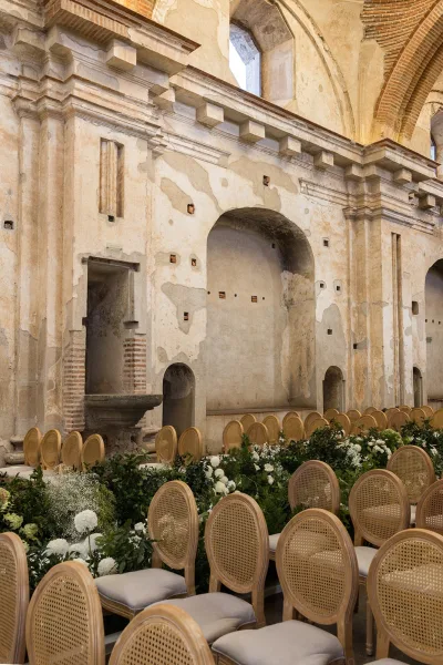 Indoor ceremony setup with wedding ceremony aisle greenery, cane chairs, and white flowers lining an aisle in a stone-arched historic venue interior