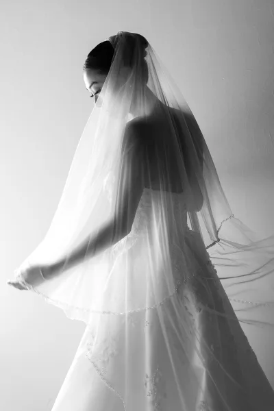 Bridal portrait in black and white of a bride looking down in a lace dress with embroidered veil trim draped over her face by a plain wall