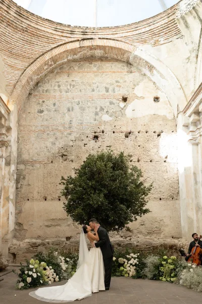 Wedding kiss portrait of bride and groom kissing under a brick arch, her veil trailing over the dress near white-green florals and a cello