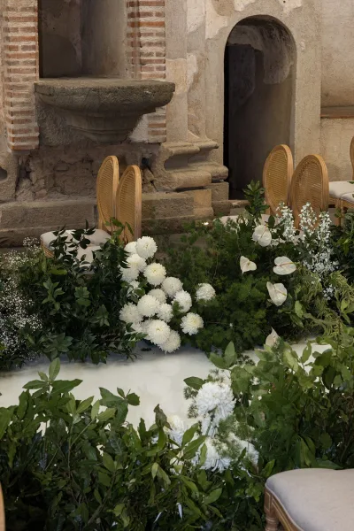 Ceremony aisle decor with a greenery aisle runner and grounded white florals, anthurium accents, and rattan chairs by a stone wall archway