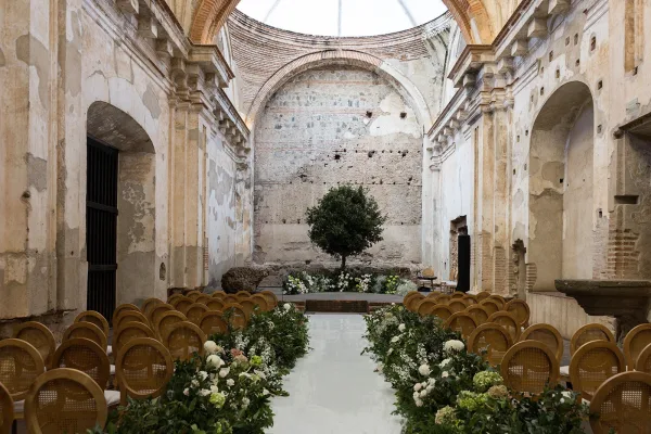 Ceremony setup with indoor wedding ceremony aisle florals and greenery garland, wooden chairs facing a tree altar in a stone-arched hall