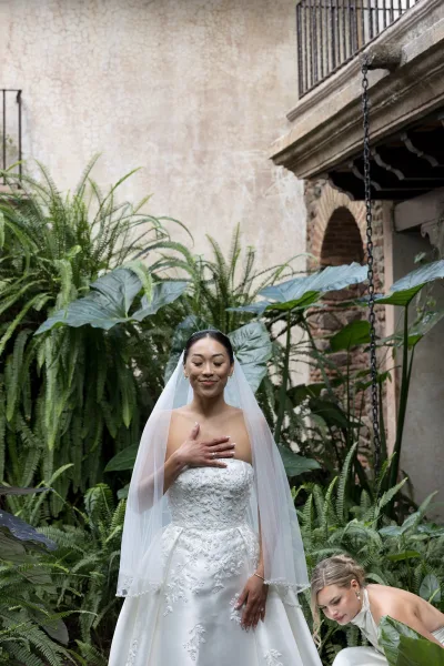 Bridal portrait of a bride in veil and strapless lace wedding dress, hand on chest showing ring, amid lush garden greenery by a stone archway