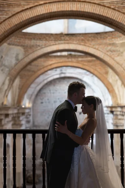 Couple portrait of bride and groom embrace with a forehead kiss, her veil and strapless dress framed by brick archways indoors