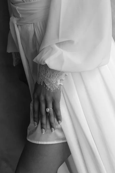 Bridal robe detail with an engagement ring close up on manicured hand, lace cuff and satin sash framed by soft fabric folds