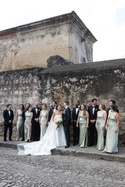 Wedding party portrait with bride holding a white bouquet beside groom, bridesmaids in sage satin and groomsmen in tuxedos by a weathered stone wall