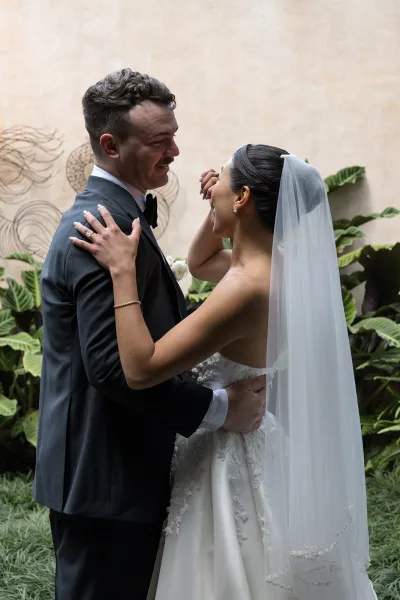 Wedding couple portrait of bride and groom embrace as she touches his face, veil flowing by a stucco wall with tropical greenery