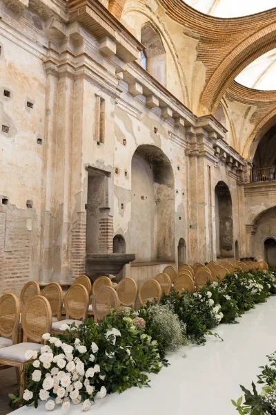 Wedding ceremony setup with a white aisle runner, greenery garland, and white roses lining the aisle inside a historic stone-arched hall