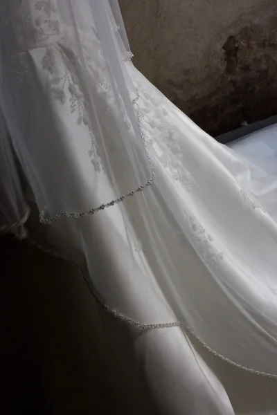 Wedding dress detail with sheer tulle overlay and beaded trim, embroidered applique and veil draped against a stone wall in shadowed interior