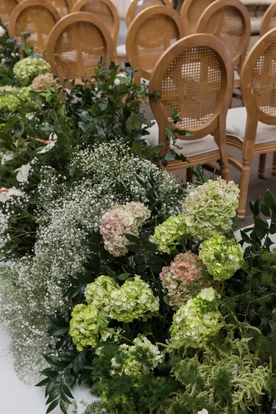 Ceremony aisle flowers with hydrangeas and baby’s breath lining a greenery garland runner beside wooden cane-back chairs on a stone floor