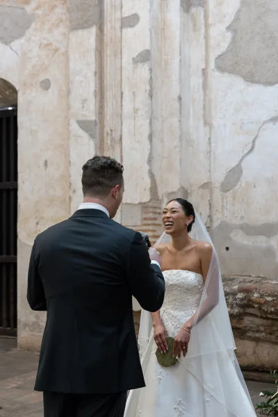Wedding vows as groom reads into a microphone while bride laughs in veil and strapless dress, framed by a brick archway courtyard