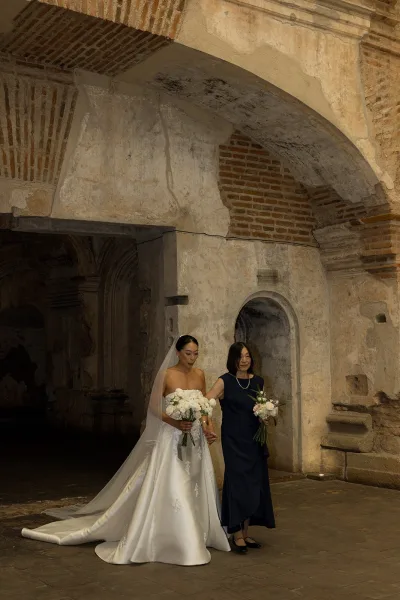 Bridal processional as bride walking down aisle with her mother, holding a white rose bouquet beneath stone arches in a historic corridor