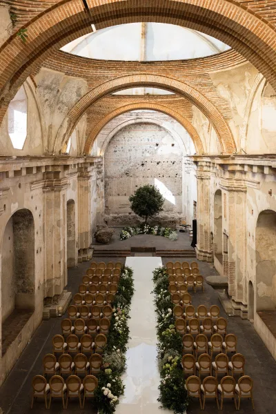 Ceremony setup with indoor wedding ceremony white aisle runner, greenery garland and floral clusters amid wooden bistro chairs under arched brick ceiling