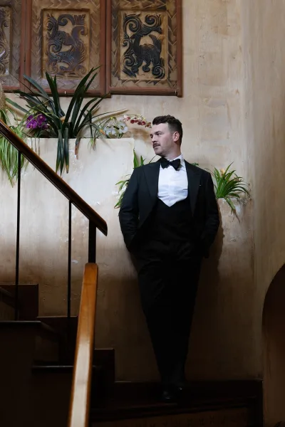 Groom portrait in a black tuxedo with boutonniere, hands in pockets on an indoor staircase beside potted orchids and carved panels